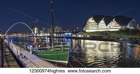 Stock Photograph - A green fishing boat moored along the edge of a river at nighttime with an illuminated Sage Gateshead in the background; Newcastle, Tyne and Wear, England. Fotosearch