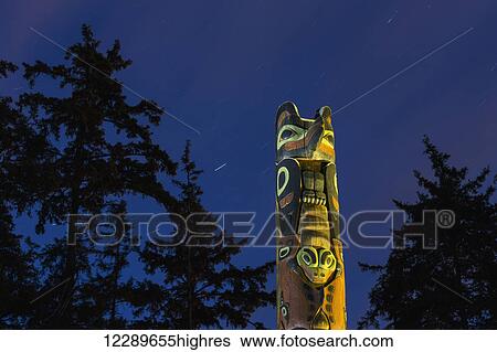 Stock Photograph - A large Totem Pole lit up at night in Sitka National Historic Park with star trails overhead, Sitka, Southeast Alaska, USA, Summer. Fotosearch