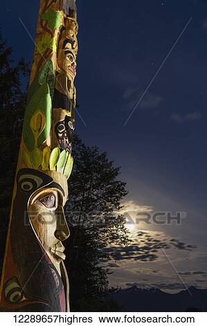Stock Photograph - A large totem pole lit up at night in Sitka National Historic Park with the moon and clouds in the background; Sitka, Alaska, United States of America. Fotosearch