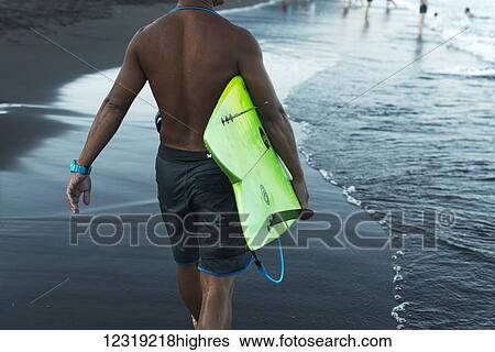 Stock Photograph - A male surfer carries his surfboard along the beach at the water's edge, Waiao beach, Yilan County; Yilan, Taiwan, China. Fotosearch
