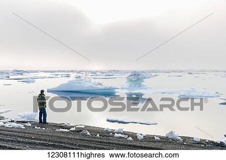 Stock Photograph - A man at the shore of the Arctic Ocean looking out towards the sun setting through dense fog, Barrow, North Slope, Arctic Alaska, USA, Summer. Fotosearch