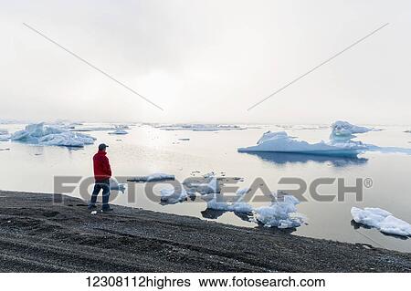 Stock Photograph - A man at the shore of the Arctic Ocean looking out towards the sun setting through dense fog, Barrow, North Slope, Arctic Alaska, USA, Summer. Fotosearch