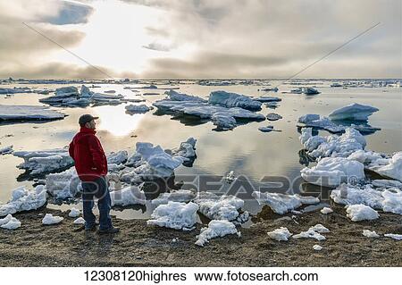 Stock Photograph - A man at the shore of the Arctic Ocean looking out towards the sun setting through dense fog, Barrow, North Slope, Arctic Alaska, USA, Summer. Fotosearch
