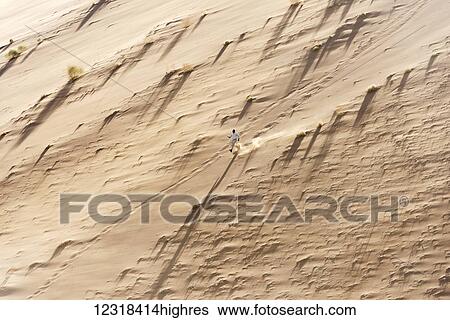 Stock Photograph - A man in modern clothes trying to climb down and running from top of a sand dune in Namib Desert; Namibia. Fotosearch