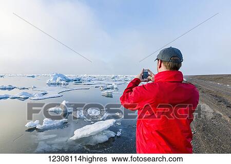 Stock Photograph - A man standing on the coast of the Arctic Ocean during a foggy sunset takes a picture with his cell phone with sea ice floating in the background; Barrow; North Slope; Arctic Alaska; USA; Summer. Fotosearch