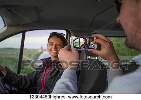 Stock Photograph - A man takes a photograph of a young woman with a smart phone while sitting in a vehicle; Egmond aan Zee, Holland. Fotosearch
