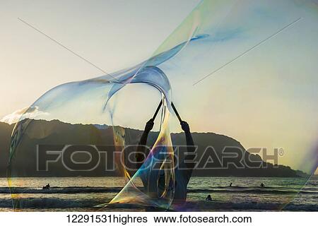 Stock Photograph - A man uses wands to create large bubbles in Hanalei Bay; Kauai, Hawaii, United States of America. Fotosearch