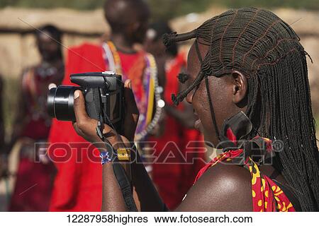 Stock Photograph - A Masai woman in traditional dress is taking pictures of other Masai while holding the camera in front of her face; Narok, Kenya. Fotosearch