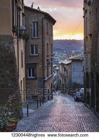 Stock Photograph - A narrow cobblestone street between buildings at sunset; Orvieto, Umbria, Italy. Fotosearch