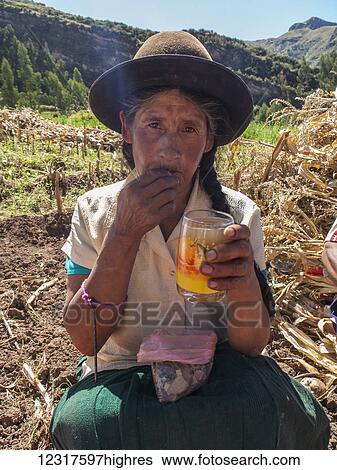 Stock Photograph - A Quechua farmer drinks chicha, a fermented corn drink and chews on Coca leaves, which are a mild stimulant; Andahuaylillas, Cusco, Peru. Fotosearch