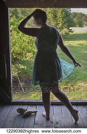 Stock Photograph - A red-headed, caucasian teenage girl wearing a turquoise dress and no shoes stands in the hay loft of an old barn framed by the open loft window overlooking empty, grass fields; Pittsboro, North Carolina, United States of America. Fotosearch