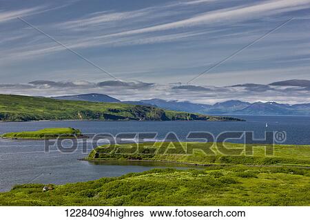 Stock Photograph - A small building sits on the coastline of Sand Point, with islands and mountians in the background, Sand Point, Southwestern Alaska, USA, Summer. Fotosearch