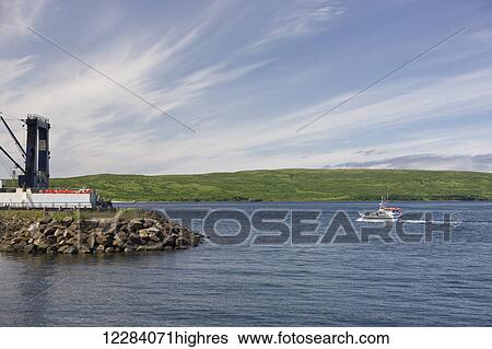 Stock Photograph - A small fishing boat motors past a breakwater in the Sand Point Harbor, Sand Point, Southwestern Alaska, USA, Summer. Fotosearch