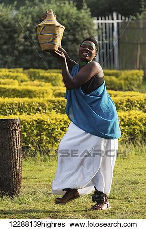 Stock Photograph - A smiling black woman in a blue top and white skirt with green bands around her neck and forehead holds up a brown pot, dancing in a garden beside a tribal drum; Nkuli, Western Province, Rwanda. Fotosearch