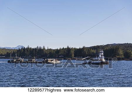 Stock Photograph - A tug boat pulls a barge out of the harbor into the ocean on a clear day on the Tongass Narrows, Ketchikan, Southeast Alaska, USA, Spring. Fotosearch