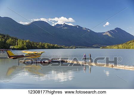 Stock Photograph - A woman and a man stand on a dock where a float plane is docked, Trail Lake Float Plane Base, Moose Pass, Kenai Peninsula, Southcentral Alaska, USA. Fotosearch