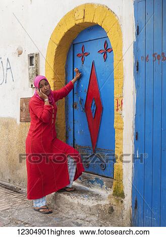A woman in a pink headscarf and red dress stands at the colourful door of a house talking on her cell phone; Essaouira, Morocco View Large Photo Image Stock Photograph - A woman in a pink headscarf and red dress stands at the colourful door of a house talking on her cell phone; Essaouira, Morocco. Fotosearch