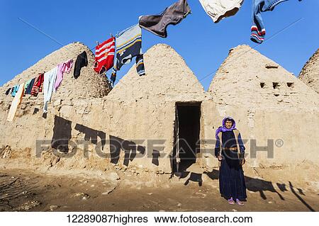 Stock Photograph - A woman standing outside the doorway of her primitive dwelling in the shadow of clothing hanging on the clothesline; Harran, Turkey. Fotosearch