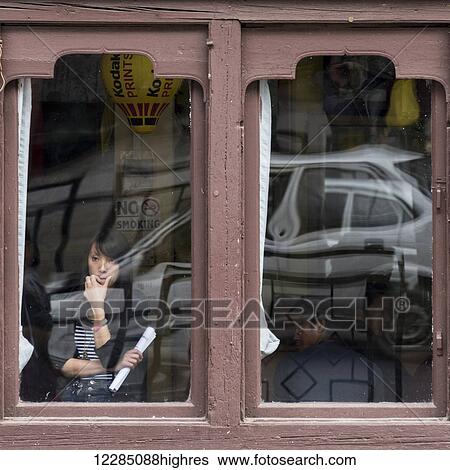 Stock Photograph - A woman stands looking out the window with a car and street reflected in the window; Thimphu, Bhutan. Fotosearch