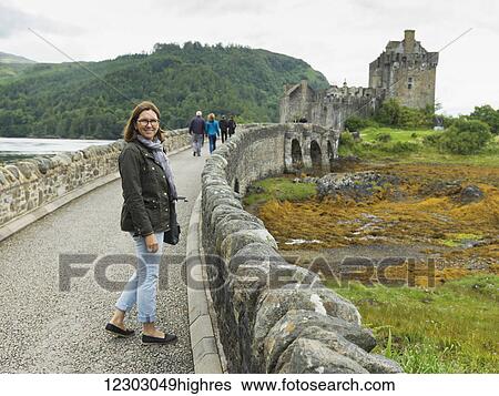 Stock Photograph - A woman stands on a stone bridge with Eilean Donan castle in the distance; Scotland. Fotosearch