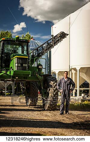 Stock Photograph - A young farmer stands posing beside a large tractor and grain storage bins on his farm; Herschel, Saskatchewan, Canada. Fotosearch