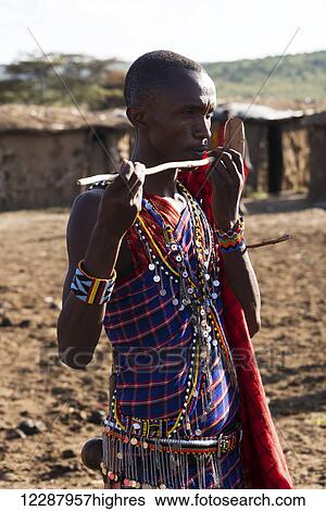 Stock Photograph - A young Masai man dressed in a traditional costume of blue, black and red checked clothing pretends to play the flute with a stick in the middle of his village; Narok, Kenya. Fotosearch