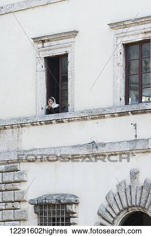 Stock Photograph - A young woman with a headscarf looks out the window of a white building; Montepulciano, Toscana, Italy. Fotosearch
