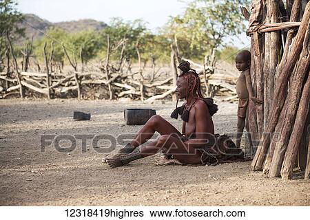 Stock Photograph - Aged woman from Himba tribe in traditional clothes and haircut is sitting near the wooden hut wall at Kunene Region; Namibia. Fotosearch