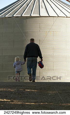 Stock Photograph - Agriculture - A farmer and his young daughter walk hand-in-hand through his farm yard with a grain bin in the background / near Sioux City, Iowa, USA.. Fotosearch