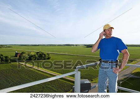 Stock Photograph - Agriculture - A young farmer talks on his cell phone atop a grain bin with a farmstead and fields in the background / near Northrop, Minnesota, USA.. Fotosearch