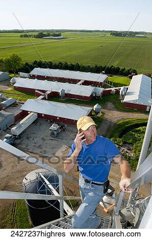 Agriculture - A young farmer talks on his cell phone atop a grain bin with a farmstead and fields in the background / near Northrop, Minnesota, USA. View Large Photo Image Stock Photograph - Agriculture - A young farmer talks on his cell phone atop a grain bin with a farmstead and fields in the background / near Northrop, Minnesota, USA.. Fotosearch