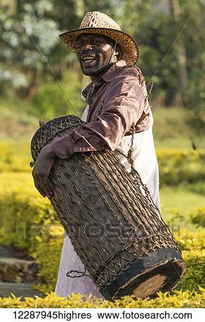 Stock Photograph - An African man in a brown shirt and pink and white patterned wrap with a leopard-print hat plays a drum; Nkuli, Western Province, Rwanda. Fotosearch