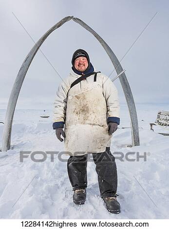 Stock Photograph - An Alaska native hunter wearing a white Kuspuk stands underneath the Browerville whalebone arch, Barrow, North Slope, Arctic Alaska, USA, Winter. Fotosearch