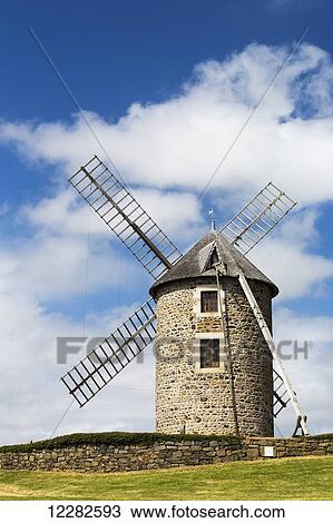 Stock Image - An old stone windmill on a hillside with wooden blades, surrounded by a stone fence with blue sky and clouds; Brehec, Brittany, France. Fotosearch