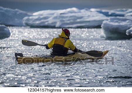 Stock Photograph - Backlit kayaker paddling by icebergs in sunlight; Antarctica. Fotosearch