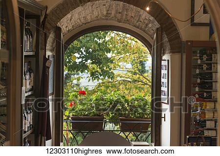 Stock Photograph - Bottles of wine on shelves in a wine shop with a view of trees and plants out the open doorway; Gaiole in Chianti, Toscana, Italy. Fotosearch