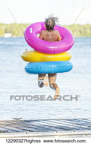 Stock Photograph - Boy jumping off of dock by Balsam Lake wearing colorful inflatable rings; Ontario, Canada. Fotosearch