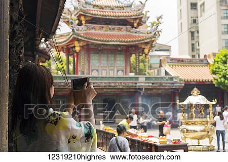 Stock Photograph - Buddhist Longshan temple in the city center of Taipei, people praying and burning incense while a tourist take a picture; Taipei, Taiwan, China. Fotosearch