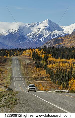 Stock Photograph - Car drives on the Alaska Highway with snow capped peaks of the St. Elias mountains and colorful trees in the background, Yukon Territory, Canada, autumn. Fotosearch