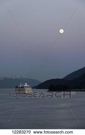 Stock Image - Celebrity cruise ship Oceania leaves Juneau harbor as the moon rises over the Inside Passage, Southeast Alaska. Fotosearch