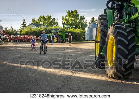 Stock Photograph - Children riding bicycles beside a large tractor on the family farm as the parents stand watching in the background; Herschel, Saskatchewan, Canada. Fotosearch