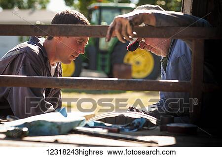 Stock Photograph - Close up of a farmer and his son sitting together on a farm beside a rail fence with a tractor in the background; Herschel, Saskatchewan, Canada. Fotosearch