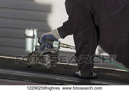 Stock Photograph - Close up of air nailer, with male roofer nailing new shingles; Calgary, Alberta, Canada. Fotosearch