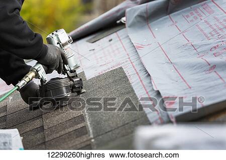 Stock Photograph - Close up of air nailer, with male roofer nailing new shingles on top of roof; Calgary, Alberta, Canada. Fotosearch
