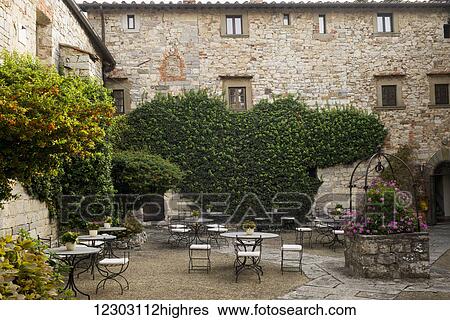Stock Photograph - Courtyard filled with tables and chairs, vines and shrubs growing around stone buildings; Gaiole in Chianti, Toscana, Italy. Fotosearch