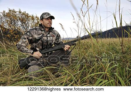 Stock Photograph - Duck hunter waits in tall grass with a shotgun in the Chugach Mountains of South-central Alaska, Autumn. Fotosearch