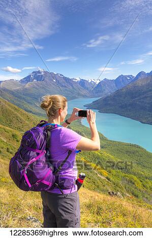 Stock Photograph - Female hiker using a smartphone to photograph Eklutna Lake and the Chugach Mountains from the Twin Peaks Trail, Chugach State Park, Southcentral Alaska, Summer. Fotosearch