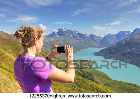 Stock Photograph - Female hiker using a smartphone to photograph Eklutna Lake and the Chugach Mountains from the Twin Peaks Trail, Chugach State Park, Southcentral Alaska, Summer. Fotosearch