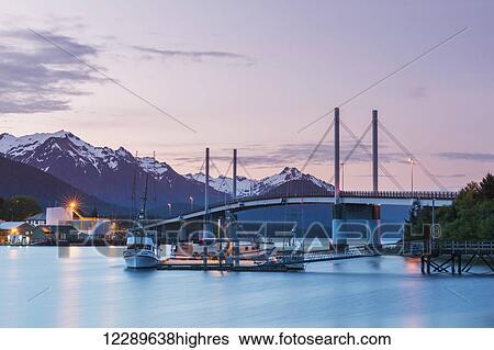Stock Photograph - Fishing boat moored along the pier in Sitka Harbor with O'Connell Bridge and the mountains in the background at twilight, Southeast Alaska, USA, Summer. Fotosearch