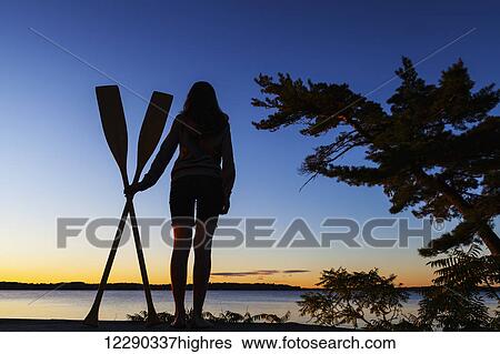 Stock Photograph - Girl holding paddles overlooking Balsam Lake at sunrise; Ontario, Canada. Fotosearch